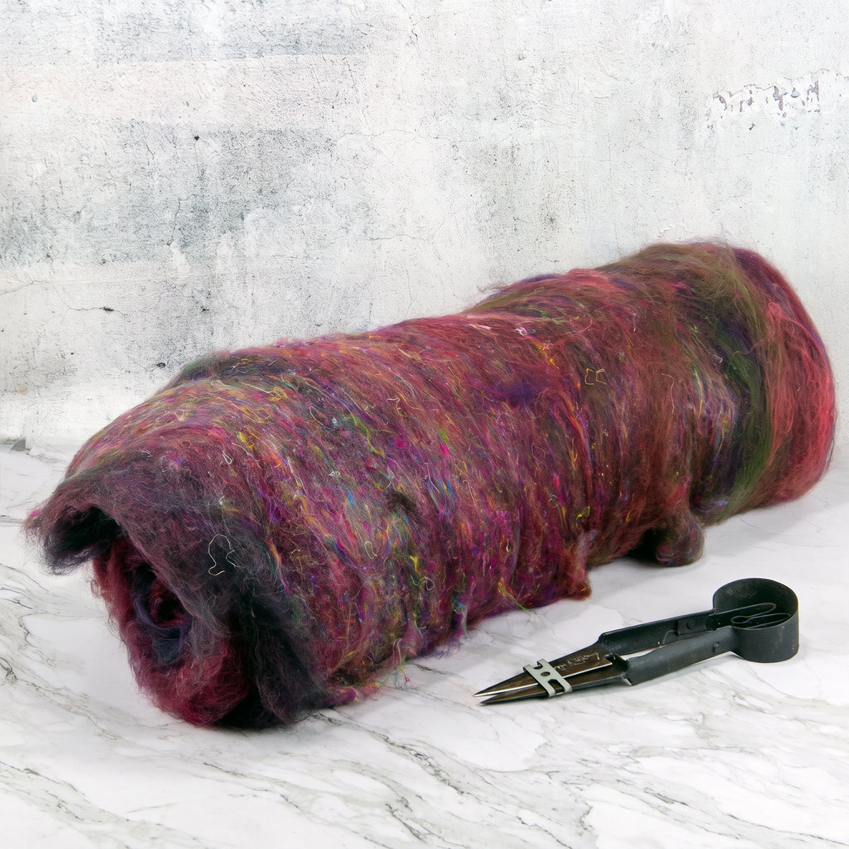 A bundle of fluffy wool roving in mixed burgundy, purple, pink, and green tones, photographed on a light stone surface with a pair of black shears beside it.