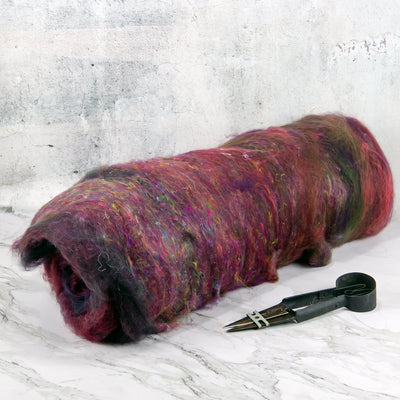 A bundle of fluffy wool roving in mixed burgundy, purple, pink, and green tones, photographed on a light stone surface with a pair of black shears beside it.